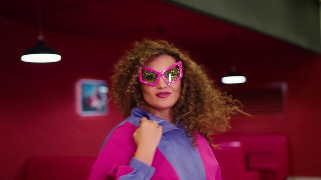 Vibrant red room with pink couches and hanging lights. Woman adjusts pink glasses, candy stick in mouth. Curly hair frames confident expression. Background features pink couches and TV screens