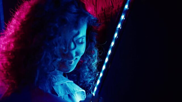 Woman holds a red feather boa. She wears a denim jacket and smiles confidently. Blue and pink LED lights illuminate the scene. The dim club setting emphasizes vibrant lighting