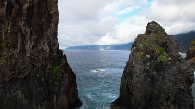 Ribeira da Janela Madeira drone backward flight emerging between dramatic sea stack rocks, revealing wild pebbled beach, rugged mountain coast and stormy sky