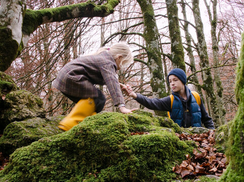 Two children explore a rocky area in the forest, one child climbs on moss-covered stones while the other assists. autumn foliage is visible on the trees