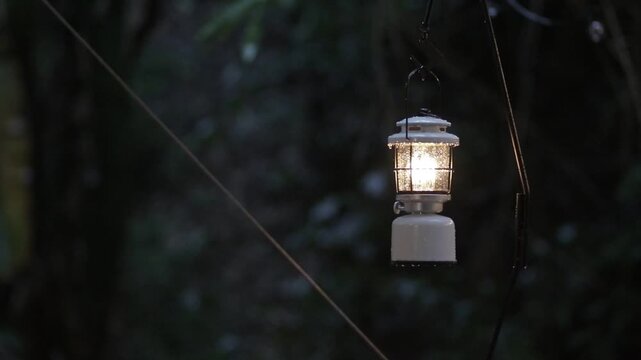 Vintage white camping lantern hanging and glowing warm light during rain at night. Cozy and peaceful outdoor campsite atmosphere.