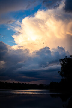 A soaring thunderhead glows with late light above a peaceful river while dark flanking clouds drift past and the smooth surface below reflects delicate tones of evening