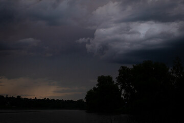 Obraz premium Dim evening light outlines dense storm layers above calm water while distant trees are barely visible and the scene feels hushed reflective and charged with the memory of rain
