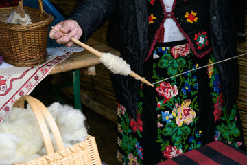 Torlak wool spinning detail shows hands using a wooden spindle with wool thread, fleece basket, embroidered tablecloth, black vest, apron, and jacket. © Visual Voyager