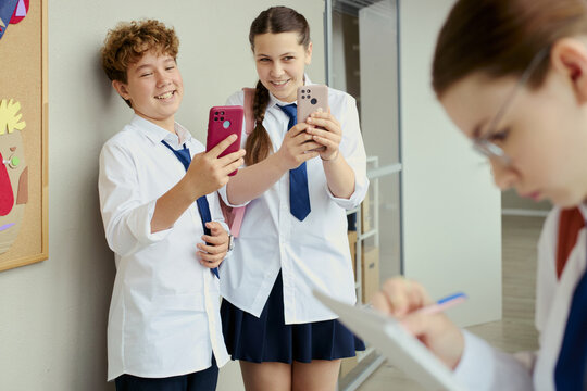 Teenage boy and teenage girl using smartphones in school hallway, smiling and sharing social media content while classmate studying nearby. Ideal for digital learning or teen communication campaigns