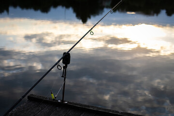Carp rig with bite alarm and bobbin stands over mirror still water. The setup watches the line while clouds drift in reflection and the angler savors the hush of a settling day. © Chornogirskyi