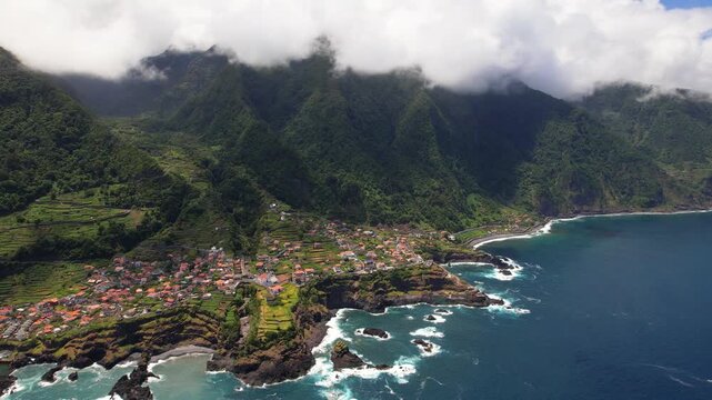 Seixal village Madeira island Portugal 4K. Aerial drone panning shot left to right of green ridged coast mountains covered in big clouds. Ocean view scenic travel b roll stock video