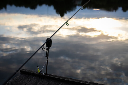 Carp rig with bite alarm and bobbin stands over mirror still water. The setup watches the line while clouds drift in reflection and the angler savors the hush of a settling day.