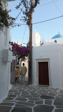 A diverse couple strolls through picturesque alleyways of Naousa, Paros, Greece, where whitewashed buildings meet vibrant bougainvillea. Experience the beauty of the Cyclades 
