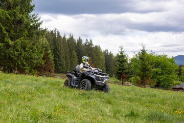 Helmeted ATV Rider Crossing a Grassy Mountain Meadow. Helmeted rider driving an ATV across a grassy meadow beside an evergreen forest under a cloudy sky. © Iryna