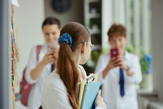 Teenage schoolgirl holding notebooks while classmates photographing her with smartphones in hallway, showing bullying and peer harassment. For school safety, teen mental health campaigns