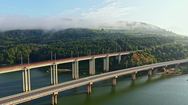 Off-center aerial perspective reveals two parallel multi-lane bridges crossing calm river, with steady traffic on upper span, lighter movement below, and forested hills fading into soft mist