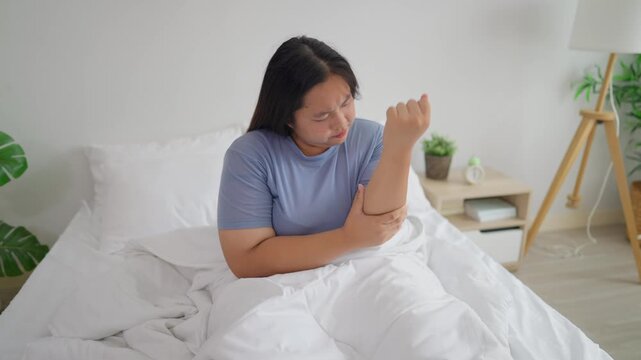 Asian plus-size woman sit on bed in morning, holding her aching hand. 