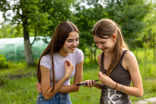 Two Teenage Friends Sharing a Smartphone Outdoors. Two smiling teenage girls looking at a smartphone together in a green garden.