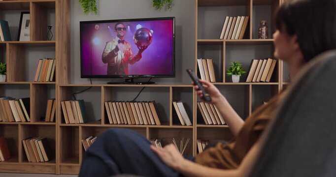 Woman watching television at home with remote. She is relaxing on the sofa enjoying a show in a living room with shelves, reflecting modern lifestyle. Simple home tv entertainment concept.