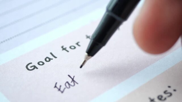 Person writing eat healthy food as goal for today in personal planner with black pen on white paper