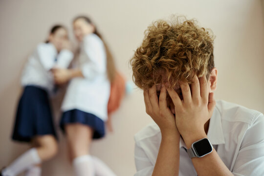 Teenage boy covering face while experiencing school bullying, classmates whispering in background. Concept for teen mental health, peer pressure, counseling campaigns, close up