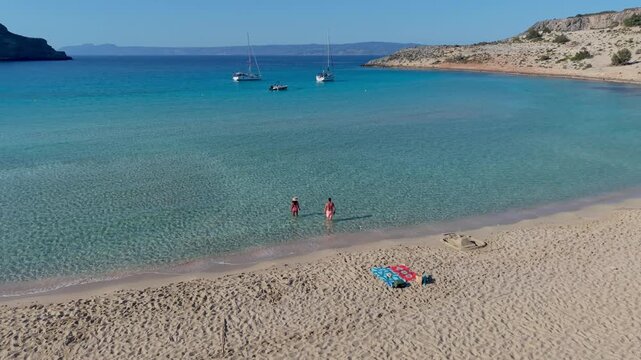 Explore Simos Beachs stunning turquoise waters and golden sands in Elafonisos, Greece. A couple relax on the beach and enjoy the serene beauty of this picturesque location while sailing boats glide by
