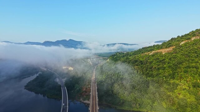 High oblique aerial view shows train moving along long railway bridge over calm river as low fog drifts through valley and clears, revealing sunlit green hillside and distant mountains