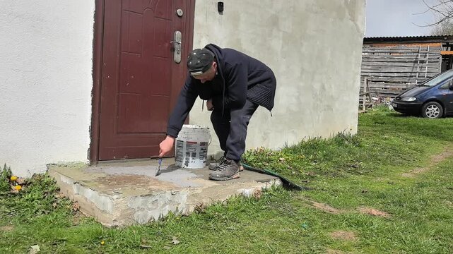 A person is applying paint to a concrete slab outside a house. The individual kneels on the ground and carefully uses a brush to cover the surface. The house has a simple design with a wooden door, 