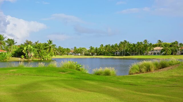 A short-cut lawn in a tropical golf course. Green lawn and beautiful view of the sky over the lake with palm trees on a sunny day. Golf course landscape. Concept of sports and outdoor lifestyle.