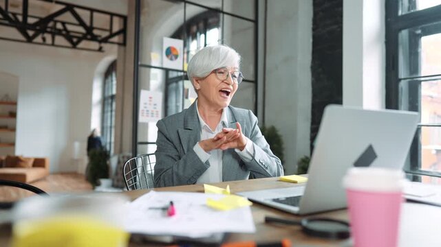 Portrait of successful mature businesswoman in modern office, smiling lady. Funny senior woman looking at laptop screen and rejoicing, investments and startup, dancing accountant or financial director