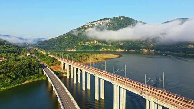 High vantage shows elevated bridge over calm river with vehicle flow toward green hills under soft mist and warm sunrise light parallel roadway and shoreline add depth and scale, with bridge, river