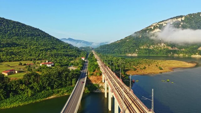Train moving across elevated railway bridge over river toward distant valley, surrounded by green hills, clear blue sky, low fog beside water, scenic daytime countryside view, with train, bridge