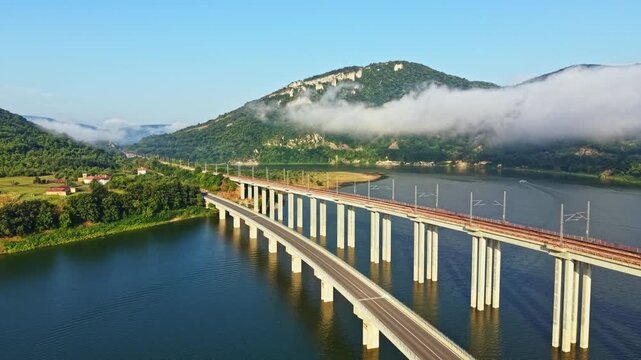 Long elevated concrete bridge stretches across calm river toward green hill wrapped in low cloud, with strong linear perspective, repeating supports, tranquil water, rural scenery and bright daylight