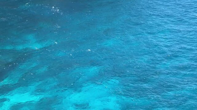 Aerial view of clear blue ocean water with varying shades of turquoise and deep blue. Sunlight reflects on the surface, creating a shimmering effect.