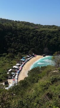 Aerial view of a secluded beach surrounded by lush green hills. The sandy shore features colorful umbrellas and beach chairs. Clear blue water gently laps at the sand.