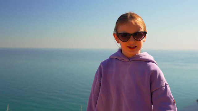 Vibrant preteen girl performs energetic hand gestures against coastal backdrop. Medium handheld tracking shot capturing rhythmic movement and sunny outdoor atmosphere.