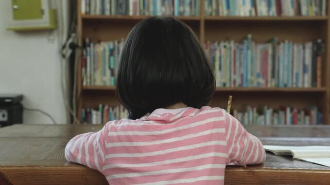 책장이 가득 찬 도서관이나 교실에서 아이가 책상에 앉아 공부하는 뒷모습입니다.