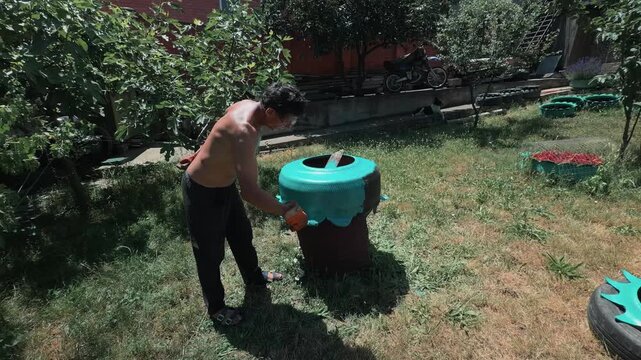 Shirtless mature Caucasian man with glasses painting upcycled scalloped car tire planter in lush backyard garden on a sunny summer day using small roller in authentic handheld camera arc shot
