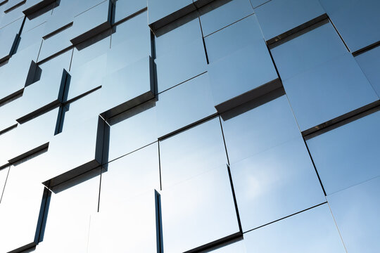 Glass facade of a contemporary commercial building featuring repeating geometric windows and reflective panels that mirror the blue sky, creating an abstract urban pattern and minimalist architecture