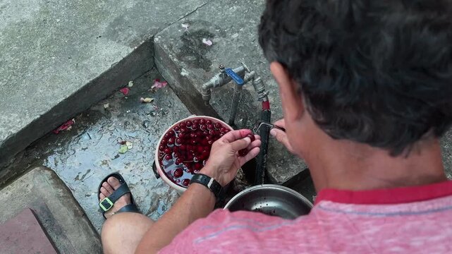 Anonymous male householder extracting pits from ripe red cherries with metal tool outdoors sitting by water tap top down over the shoulder handheld shot of authentic rural domestic harvesting chores