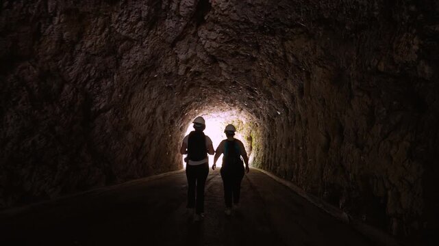 Hikers wearing safety helmets walk through the historic stone tunnel of Vecchia Strada della Valcellina.