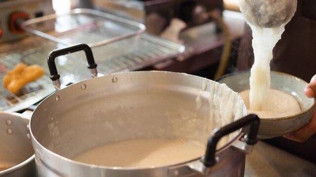 Chef Ladling Hot Rice Congee into Bowl from Large Metal Pot