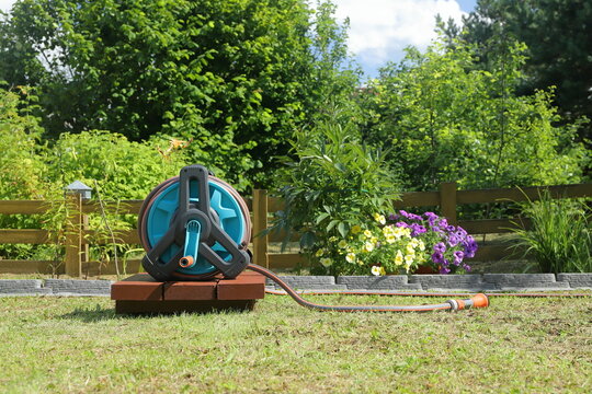 A garden hose reel with a sprayer nozzle on it stands on a wooden pedestal in front of a yellow wooden hedge in a summer garden