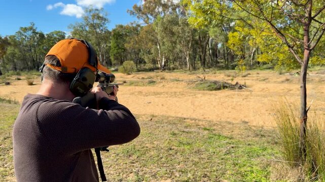 Man Shooting Scoped Rifle with Ear Protection in Sunny Forest Clearing