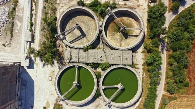 Workers at a water treatment facility are monitoring the treatment process in large round tanks. The facility is located in a rural area surrounded by trees and fields.