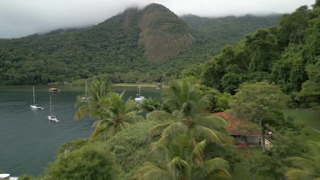 Flight over the hill with trees towards the boats anchored in a calm bay. Ilha Grande, Brazil