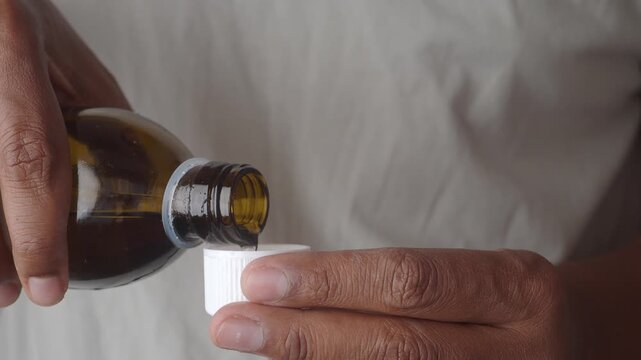 A close-up shot of a person pouring cough syrup from a dark glass bottle into a small measuring cup, illustrating medication dosage