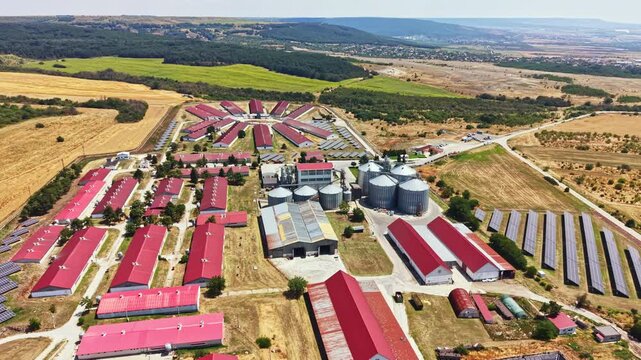 A pig farm features several structures arranged in a layout with fields and silos in the background. This farm is situated in a rural area with clear skies and open space.