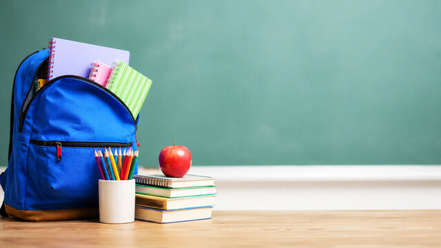 Blue backpack with school supply and red apple on wooden desk in front of green chalkboard. Education concept with student bag and stationery. Classroom background for back to school time.
