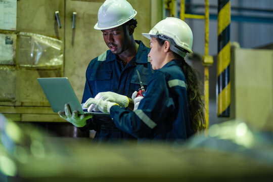 Factory technicians review maintenance records on laptop while inspecting industrial equipment together. Engineering support, production monitoring, manufacturing technology.