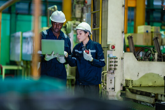 Factory engineer and technician review production data on laptop beside industrial machine while discussing maintenance tasks. Manufacturing teamwork, equipment inspection, operational efficiency.