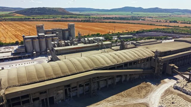 A large industrial site stands in a rural area. The building has multiple sections and is surrounded by fields and mountains. Machinery is present, indicating active production.
