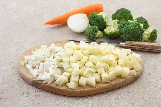 Fresh diced potatoes and chopped onion on a wooden cutting board with a kitchen knife, ready for homemade soup cooking on a light textured background