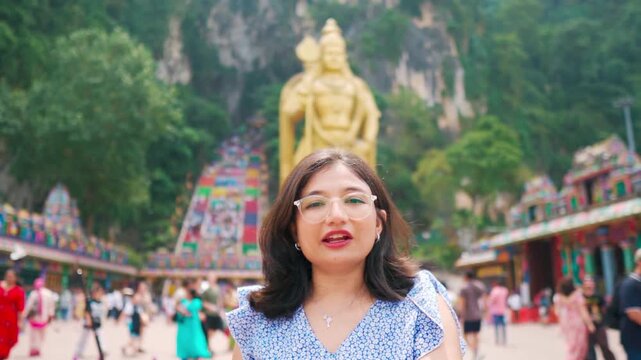 Portrait of Indian woman vlogging at Batu Caves, Kuala Lumpur, Malaysia. Vibrant temple steps and golden statue of Lord Murugan in background. Confident influencer sharing authentic travel experience.